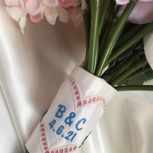 Puede incluir: Un envoltorio de ramo de satén blanco con bordados en rosa y azul. El bordado incluye las iniciales "B&C" y la fecha "4.6.21".