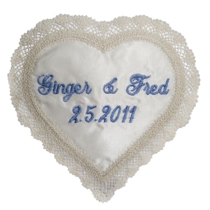 May include: White satin heart-shaped pillow with a lace trim. The pillow is embroidered with the names "Ginger & Fred" and the date "2.5.2011" in blue thread.
