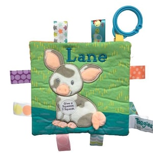 May include: A green and blue fabric baby toy with a pig on the front. The pig is white with black spots and has a pink nose. The toy has a variety of colorful fabric tags attached to it. The toy says "Give a Squeeze, I Squeak" and "Lane" on the front.