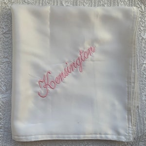 May include: White satin baby blanket with pink embroidery that says "Kensington".