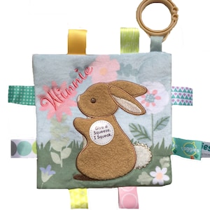 May include: A blue and green baby security blanket with a brown bunny embroidered on it. The blanket has a variety of colorful ribbons attached to it and says "Winnie" and "Give a Squeeze, I Squeak." on it.