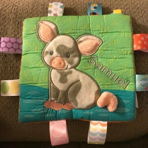 May include: A green and blue baby blanket with a pig embroidered on it. The blanket has several colorful fabric tags attached to it. The pig is gray and white with a pink nose and ears. The blanket is personalized with the name "Samuel".