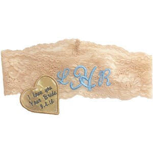 Op de afbeelding: Een beige kanten kousenband met blauwe geborduurde initialen "L&R" en een gouden hartvormige patch met de tekst "I love you Your Bride 9.4.16".