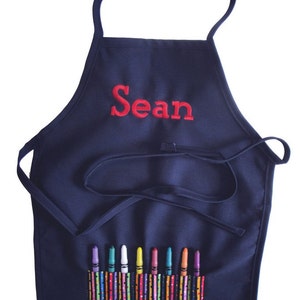 May include: A navy blue child's art smock with a pocket for crayons. The smock has the name "Sean" embroidered on the front in red thread.