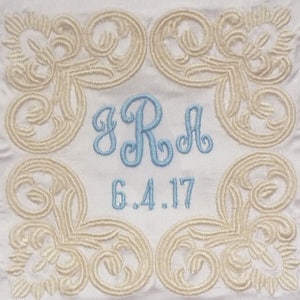 May include: White fabric with a light blue embroidered monogram "SRA" and the date "6.4.17" surrounded by a decorative border of cream-colored embroidery.