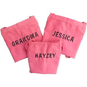 Puede incluir: Tres delantales rosas con nombres bordados en negro: Abuela, Jessica y Hayley.