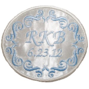 May include: White satin round patch with blue embroidered scrollwork border. The initials "RKB" and the date "6.23.12" are embroidered in the center.