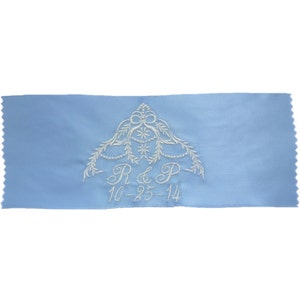 May include: A light blue satin ribbon with white embroidery. The embroidery features a decorative design with a bow at the top and the initials "R & P" and the date "10-25-14" below.