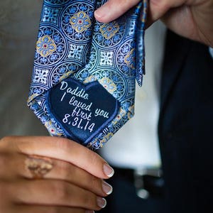 May include: A blue patterned tie with a decorative design. The tie has a navy blue patch with the embroidered text "Daddio I loved you first 8.31.14". The tie is held by a person.