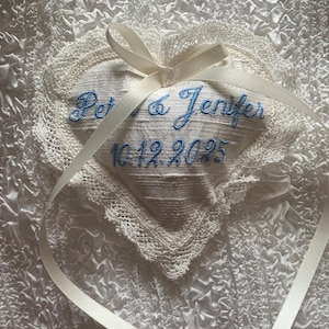 May include: Heart-shaped ring pillow with embroidered names and date in blue thread. The pillow is white with a lace trim and a cream-colored ribbon bow. The text reads "Pet & Jenifer 10.12.2025".