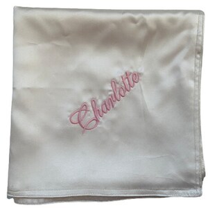 May include: A white satin baby blanket with the name "Charlotte" embroidered in pink.