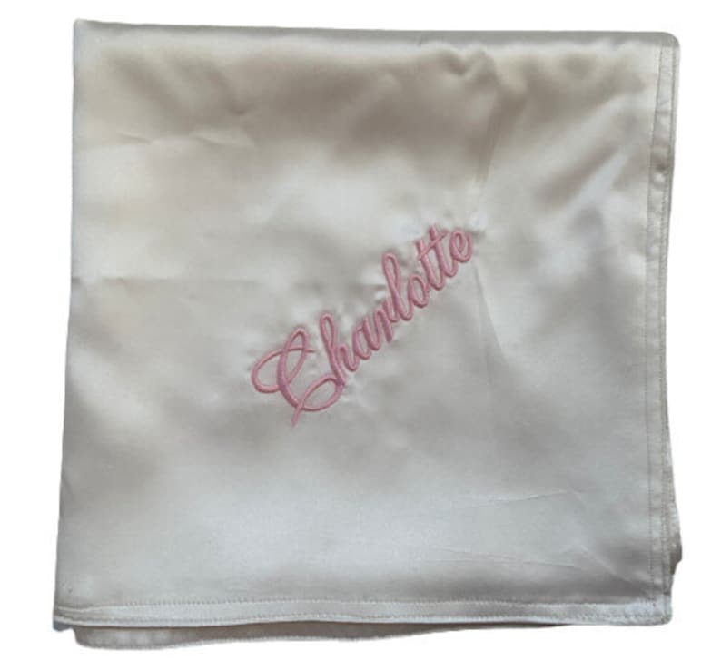May include: A white satin baby blanket with the name "Charlotte" embroidered in pink.