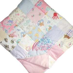 Può includere: Una trapunta per neonati patchwork con un retro rosa e un pannello centrale blu chiaro. La trapunta è realizzata con diversi tessuti con motivi diversi, tra cui motivi floreali, animali e geometrici. Il nome "Maia" è ricamato in rosa sul pannello centrale.