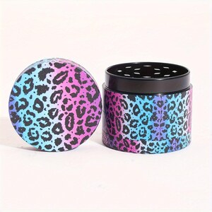 Op de afbeelding: Een driedelige grinder met een luipaardprint in roze, blauwe en paarse tinten. De grinder heeft een zwart metalen bovenkant met een zeef.