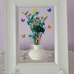 K&ouml;nnte beinhalten: Gerahmtes Blumenarrangement mit einer wei&szlig;en Vase, die getrocknete teal- und wei&szlig;farbene Blumen enth&auml;lt. Bunte Schmetterlingsakzente in Gelb, Blau, Orange und Lila schm&uuml;cken das Arrangement. Der wei&szlig;e Rahmen wird von einem transparenten St&auml;nder getragen.