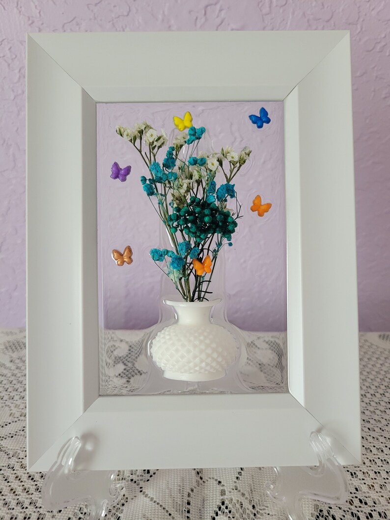 K&ouml;nnte beinhalten: Gerahmtes Blumenarrangement mit einer wei&szlig;en Vase, die getrocknete teal- und wei&szlig;farbene Blumen enth&auml;lt. Bunte Schmetterlingsakzente in Gelb, Blau, Orange und Lila schm&uuml;cken das Arrangement. Der wei&szlig;e Rahmen wird von einem transparenten St&auml;nder getragen.