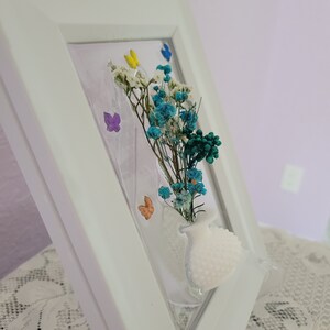 K&ouml;nnte beinhalten: Ein wei&szlig; gerahmtes Kunstwerk mit einer kleinen wei&szlig;en Vase mit getrockneten blauen, wei&szlig;en und gr&uuml;nen Blumen. Bunte Schmetterlingsakzente sind ebenfalls im Design enthalten. Der Rahmen ist abgewinkelt und zeigt die Tiefe des Kunstwerks.