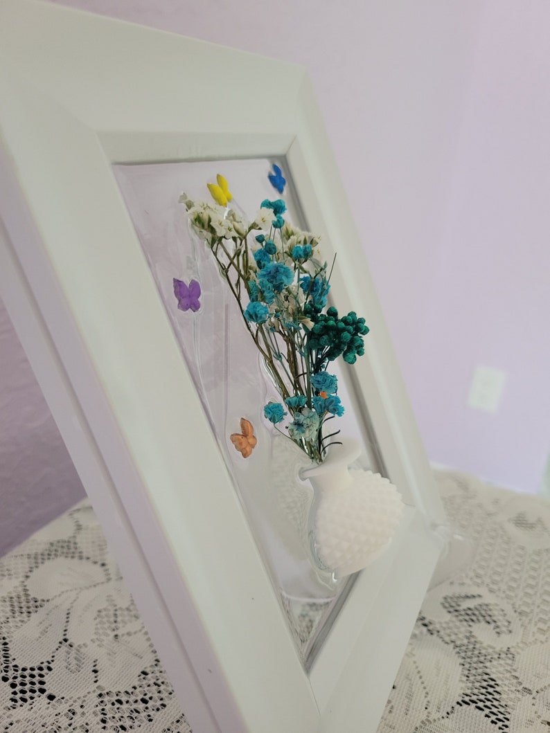 K&ouml;nnte beinhalten: Ein wei&szlig; gerahmtes Kunstwerk mit einer kleinen wei&szlig;en Vase mit getrockneten blauen, wei&szlig;en und gr&uuml;nen Blumen. Bunte Schmetterlingsakzente sind ebenfalls im Design enthalten. Der Rahmen ist abgewinkelt und zeigt die Tiefe des Kunstwerks.