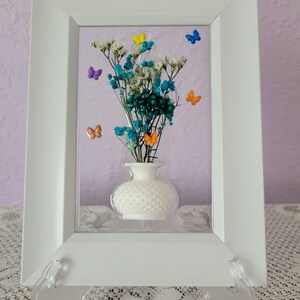 K&ouml;nnte beinhalten: Ein wei&szlig; gerahmtes Kunstwerk mit einer kleinen wei&szlig;en Vase mit getrockneten Blumen in T&uuml;rkis-, Wei&szlig;- und Gr&uuml;nt&ouml;nen. Bunte Schmetterlingsakzente in Gelb, Orange, Blau und Lila sind um die Blumen herum verstreut. Der Hintergrund ist helllila.