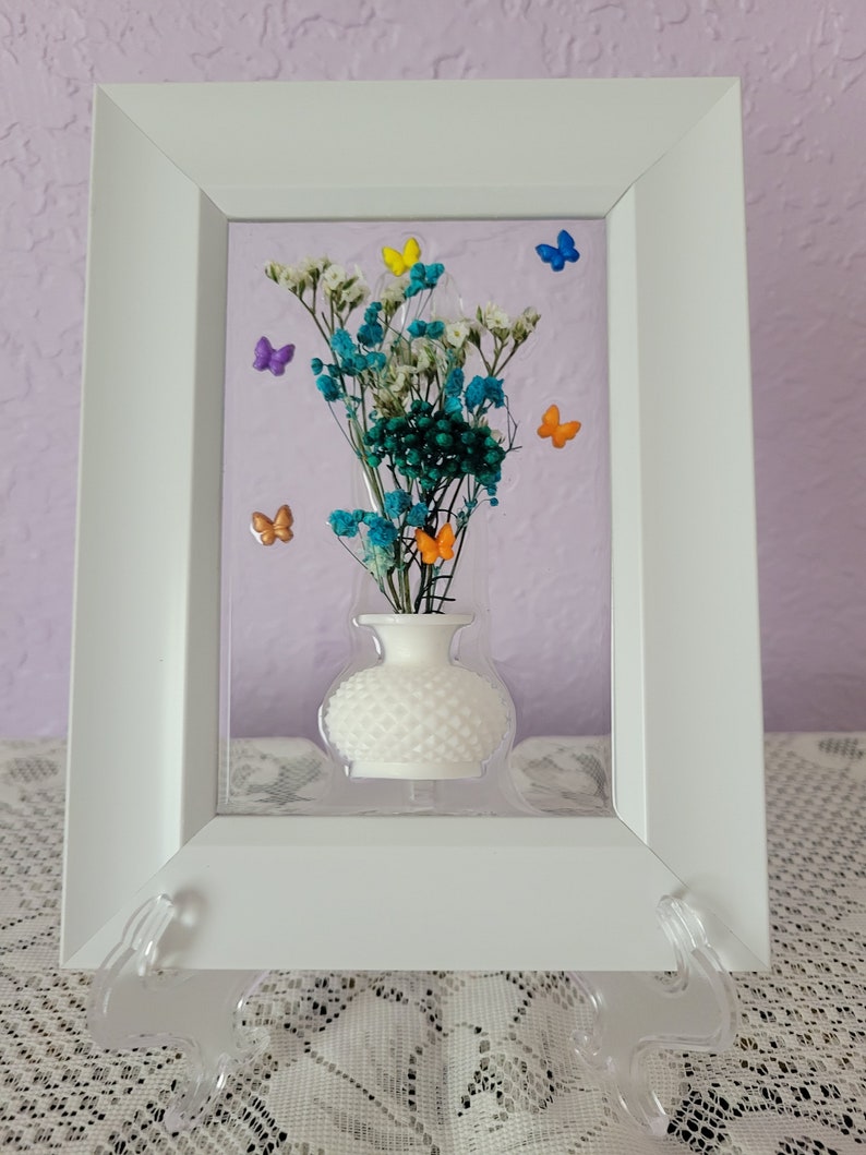 K&ouml;nnte beinhalten: Ein wei&szlig; gerahmtes Kunstwerk mit einer kleinen wei&szlig;en Vase mit getrockneten Blumen in T&uuml;rkis-, Wei&szlig;- und Gr&uuml;nt&ouml;nen. Bunte Schmetterlingsakzente in Gelb, Orange, Blau und Lila sind um die Blumen herum verstreut. Der Hintergrund ist helllila.