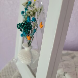K&ouml;nnte beinhalten: Ein wei&szlig; gerahmtes Kunstwerk mit einer kleinen wei&szlig;en Vase mit bunten Trockenblumen. Die Blumen sind t&uuml;rkis, wei&szlig; und orange. Der Hintergrund hat drei kleine, bunte Schmetterlinge.