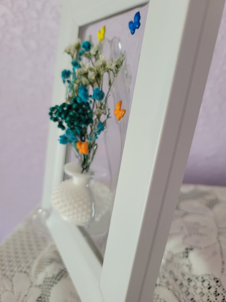 K&ouml;nnte beinhalten: Ein wei&szlig; gerahmtes Kunstwerk mit einer kleinen wei&szlig;en Vase mit bunten Trockenblumen. Die Blumen sind t&uuml;rkis, wei&szlig; und orange. Der Hintergrund hat drei kleine, bunte Schmetterlinge.