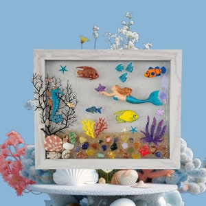Könnte beinhalten: Ein weiß gerahmtes Kunstharz-Kunstwerk, das eine Meerjungfrau zeigt, die mit Seepferdchen, Fischen und Korallen schwimmt. Der Hintergrund ist hellblau mit weißen Glitzer. Das Stück ist von weißen Muscheln umgeben.