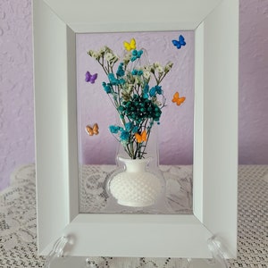May include: A white framed art piece featuring a white vase with colorful dried flowers and small butterfly accents. The flowers are teal, white, and green. The butterflies are yellow, blue, orange, and purple.