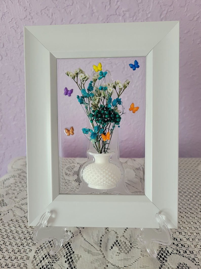 K&ouml;nnte beinhalten: Ein wei&szlig; gerahmtes Kunstwerk mit einer wei&szlig;en Vase mit bunten Trockenblumen und kleinen Schmetterlingsakzenten. Die Blumen sind t&uuml;rkis, wei&szlig; und gr&uuml;n. Die Schmetterlinge sind gelb, blau, orange und lila.