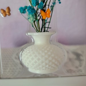 K&ouml;nnte beinhalten: Eine kleine wei&szlig;e Vase mit einem strukturierten Rautenmuster enth&auml;lt t&uuml;rkis- und orangefarbene Blumen. Zwei kleine goldene Schmetterlinge befinden sich links von der Vase. Die Vase befindet sich in einem transparenten Kunststoffbeh&auml;lter.