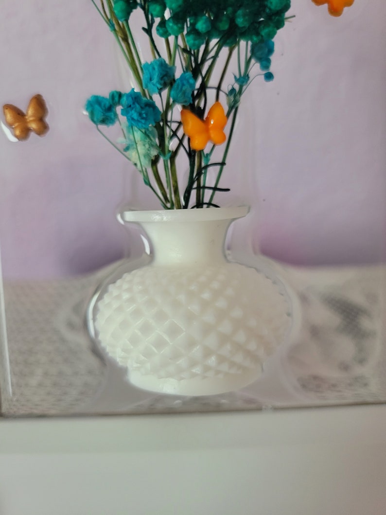 K&ouml;nnte beinhalten: Eine kleine wei&szlig;e Vase mit einem strukturierten Rautenmuster enth&auml;lt t&uuml;rkis- und orangefarbene Blumen. Zwei kleine goldene Schmetterlinge befinden sich links von der Vase. Die Vase befindet sich in einem transparenten Kunststoffbeh&auml;lter.