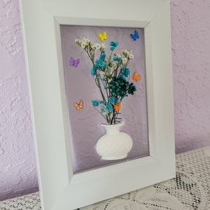 K&ouml;nnte beinhalten: Gerahmtes Blumenkunstwerk mit einer wei&szlig;en Vase mit bunten Trockenblumen und Schmetterlingsakzenten. Der wei&szlig;e Rahmen umgibt einen lavendelfarbenen Hintergrund und schafft eine dekorative Wanddekoration.