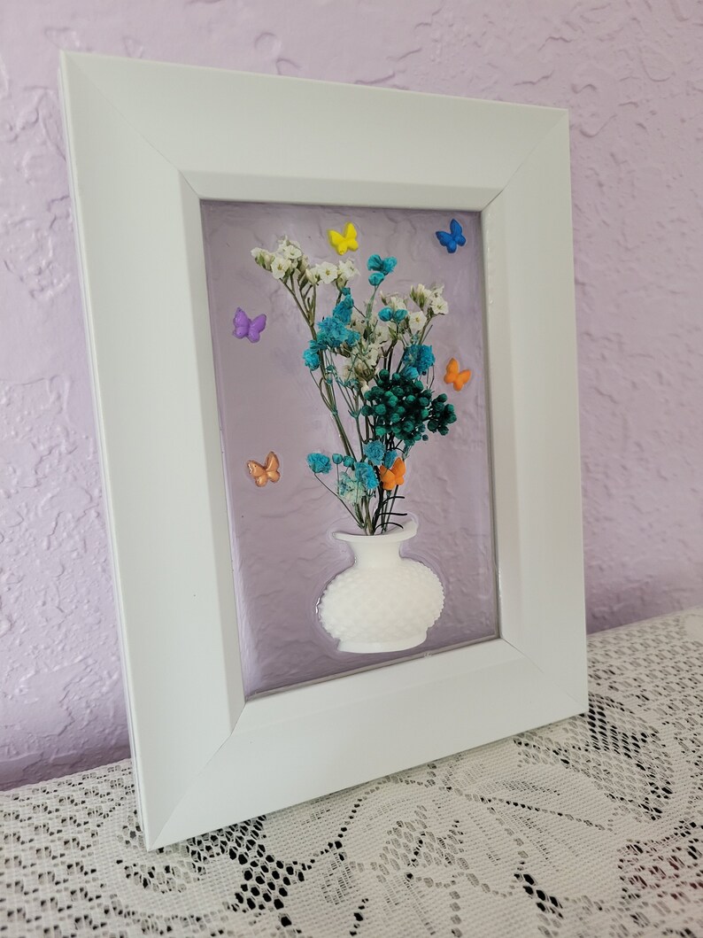 K&ouml;nnte beinhalten: Gerahmtes Blumenkunstwerk mit einer wei&szlig;en Vase mit bunten Trockenblumen und Schmetterlingsakzenten. Der wei&szlig;e Rahmen umgibt einen lavendelfarbenen Hintergrund und schafft eine dekorative Wanddekoration.