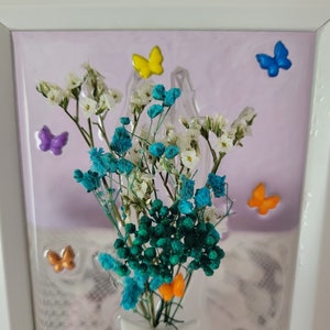 K&ouml;nnte beinhalten: Gerahmtes Blumenarrangement mit wei&szlig;en, t&uuml;rkisfarbenen und gr&uuml;nen Trockenblumen. Die Anordnung ist mit bunten Schmetterlingsverzierungen in Gelb, Blau, Orange und Lila verziert. Der Hintergrund ist helllila.