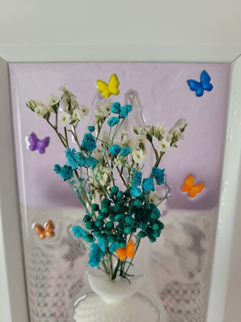 K&ouml;nnte beinhalten: Gerahmtes Blumenarrangement mit wei&szlig;en, t&uuml;rkisfarbenen und gr&uuml;nen Trockenblumen. Die Anordnung ist mit bunten Schmetterlingsverzierungen in Gelb, Blau, Orange und Lila verziert. Der Hintergrund ist helllila.