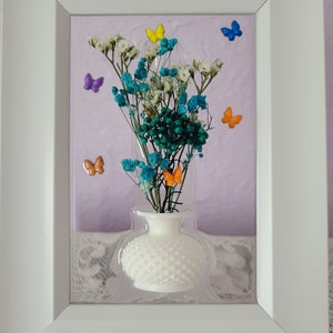 K&ouml;nnte beinhalten: Gerahmtes Blumenarrangement mit einer wei&szlig;en Vase mit strukturiertem Muster, gef&uuml;llt mit blauen, wei&szlig;en und gr&uuml;nen Trockenblumen. Bunte Schmetterlingsakzente in Gelb, Orange, Lila und Blau. Der Rahmen ist wei&szlig;.
