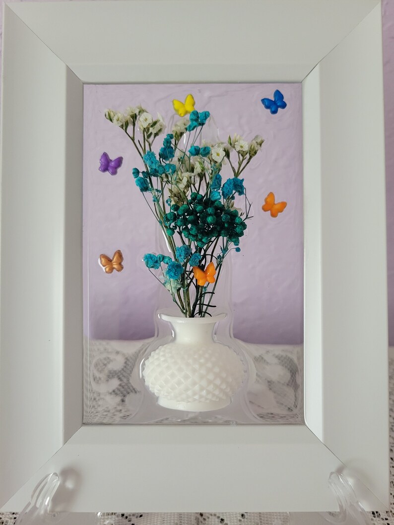 K&ouml;nnte beinhalten: Gerahmtes Blumenarrangement mit einer wei&szlig;en Vase mit strukturiertem Muster, gef&uuml;llt mit blauen, wei&szlig;en und gr&uuml;nen Trockenblumen. Bunte Schmetterlingsakzente in Gelb, Orange, Lila und Blau. Der Rahmen ist wei&szlig;.