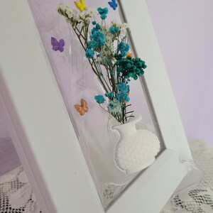 K&ouml;nnte beinhalten: Ein wei&szlig; gerahmtes Kunstwerk mit einer kleinen wei&szlig;en Vase mit einem Strau&szlig; getrockneter Blumen in Blau-, Wei&szlig;- und Gr&uuml;nt&ouml;nen. Bunte Schmetterlingsakzente sind um die Blumen herum verstreut. Der Rahmen ist abgewinkelt und zeigt die Tiefe des Kunstwerks.
