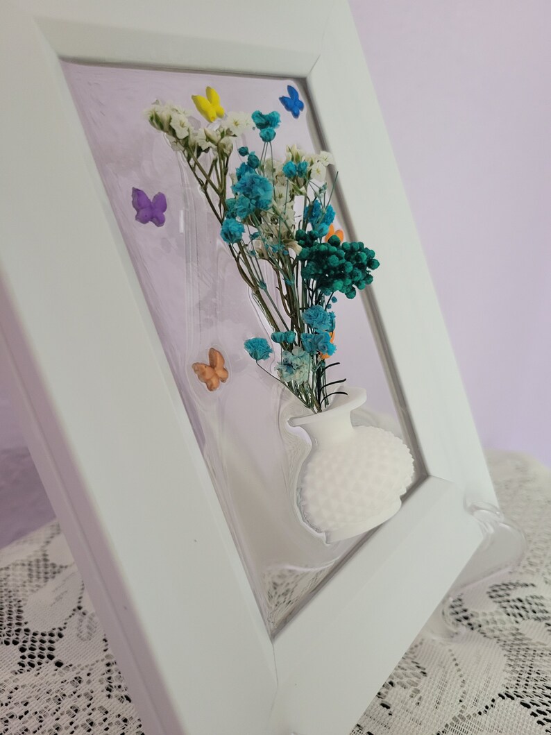 K&ouml;nnte beinhalten: Ein wei&szlig; gerahmtes Kunstwerk mit einer kleinen wei&szlig;en Vase mit einem Strau&szlig; getrockneter Blumen in Blau-, Wei&szlig;- und Gr&uuml;nt&ouml;nen. Bunte Schmetterlingsakzente sind um die Blumen herum verstreut. Der Rahmen ist abgewinkelt und zeigt die Tiefe des Kunstwerks.