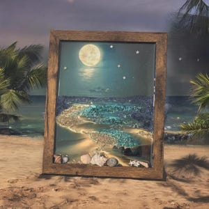 May include: A framed resin art piece depicting a moonlit beach scene with a full moon, stars, and a sandy beach with blue and green glitter. The frame is made of dark wood.