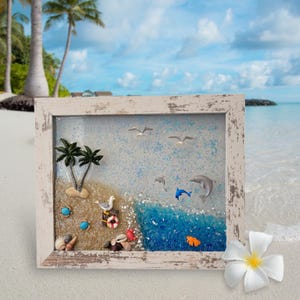 Könnte beinhalten: Ein gerahmtes Harz-Kunstwerk, das eine Strandszene mit Palmen, Möwen, Delfinen, Muscheln und einem Rettungsring darstellt. Der Rahmen ist weiß mit einer abgenutzten Oberfläche.