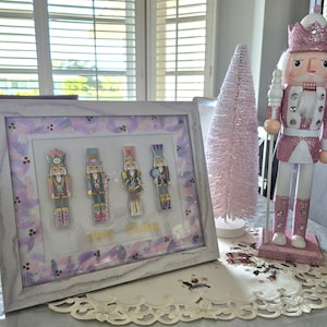 May include: A framed artwork featuring four colorful nutcracker figures with the text "Merry Christmas" in gold. A pink glittery nutcracker and a pink Christmas tree are also displayed on a table with a decorative doily.