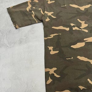 Puede incluir: Camiseta de camuflaje de manga corta en tonos verdes y beige. La camiseta tiene cuello redondo y la palabra "VEEZY" est&aacute; impresa en el cuello. La tela tiene un aspecto ligeramente desgastado.