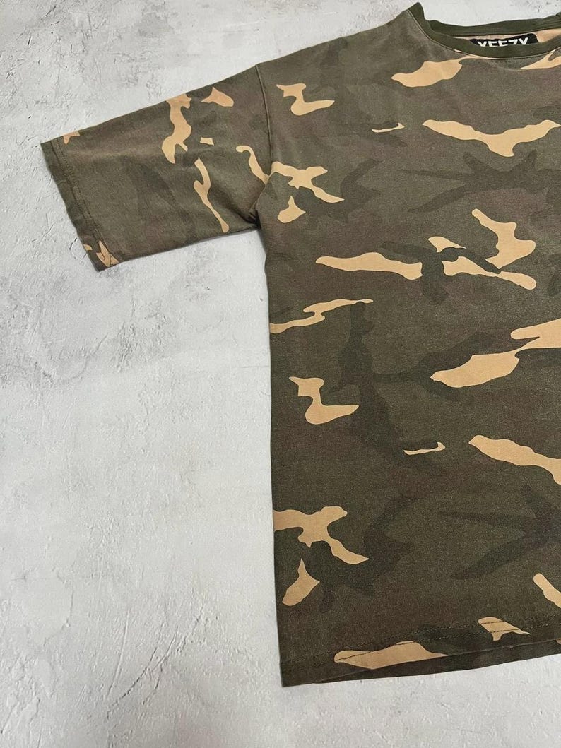 Puede incluir: Camiseta de camuflaje de manga corta en tonos verdes y beige. La camiseta tiene cuello redondo y la palabra "VEEZY" est&aacute; impresa en el cuello. La tela tiene un aspecto ligeramente desgastado.