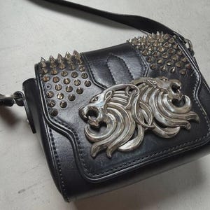Puede incluir: Bolso bandolera de cuero negro con un emblema de cabeza de le&oacute;n plateado y tachuelas. El bolso tiene un cierre de solapa y una correa ajustable. El dise&ntilde;o es atrevido y elegante, adecuado para diversas ocasiones.