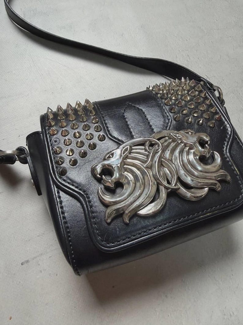 Puede incluir: Bolso bandolera de cuero negro con un emblema de cabeza de le&oacute;n plateado y tachuelas. El bolso tiene un cierre de solapa y una correa ajustable. El dise&ntilde;o es atrevido y elegante, adecuado para diversas ocasiones.