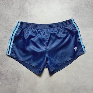 May include: Navy blue athletic shorts with light blue stripes down the sides. The shorts have an elastic waistband and a small white logo on the leg. These vintage shorts are made of a shiny, lightweight material.