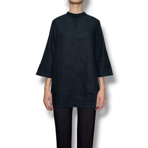 May include: A dark navy blue tunic featuring a mandarin collar, a partial button-down placket, and three-quarter length sleeves. The garment has a straight hemline and a relaxed fit, paired with solid black trousers that extend below the frame.