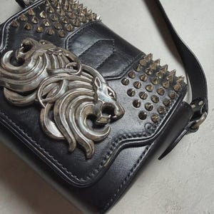 Puede incluir: Bolso bandolera de cuero negro con emblema de cabeza de le&oacute;n plateado y tachuelas. El bolso tiene un cierre de solapa y una correa larga ajustable. La parte superior y la solapa est&aacute;n adornadas con tachuelas c&oacute;nicas plateadas.