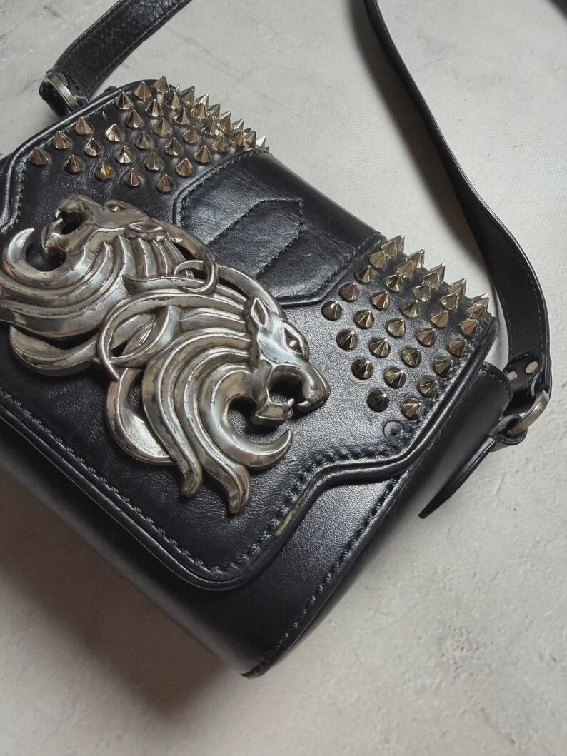 Puede incluir: Bolso bandolera de cuero negro con emblema de cabeza de le&oacute;n plateado y tachuelas. El bolso tiene un cierre de solapa y una correa larga ajustable. La parte superior y la solapa est&aacute;n adornadas con tachuelas c&oacute;nicas plateadas.
